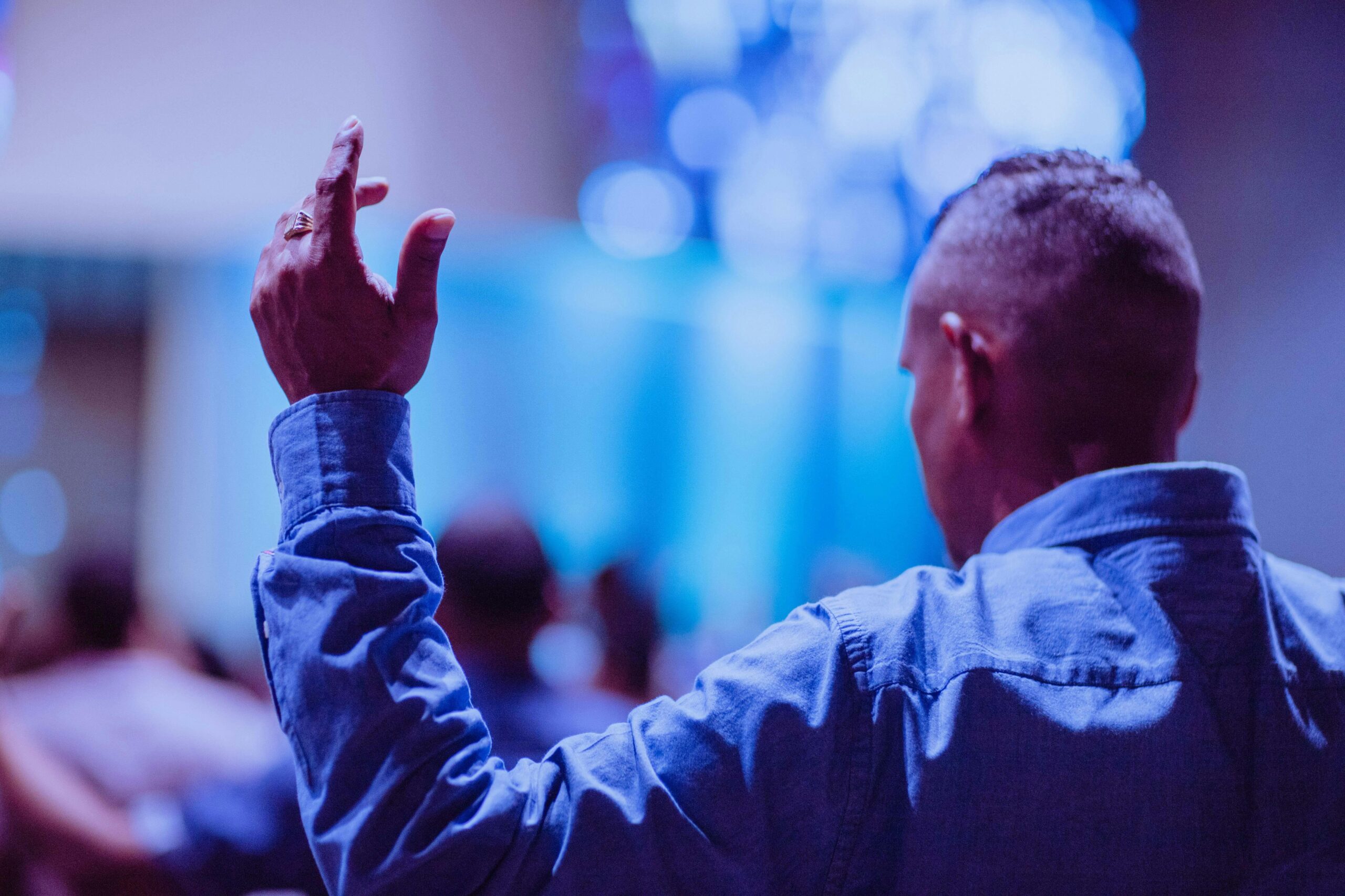 Back view of a man raising hand in prayer at a church service indoors.