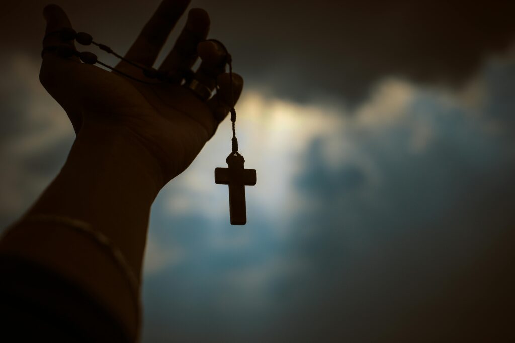 Silhouette of a hand holding a rosary with a cross against a dramatic sky, symbolizing faith and spirituality.