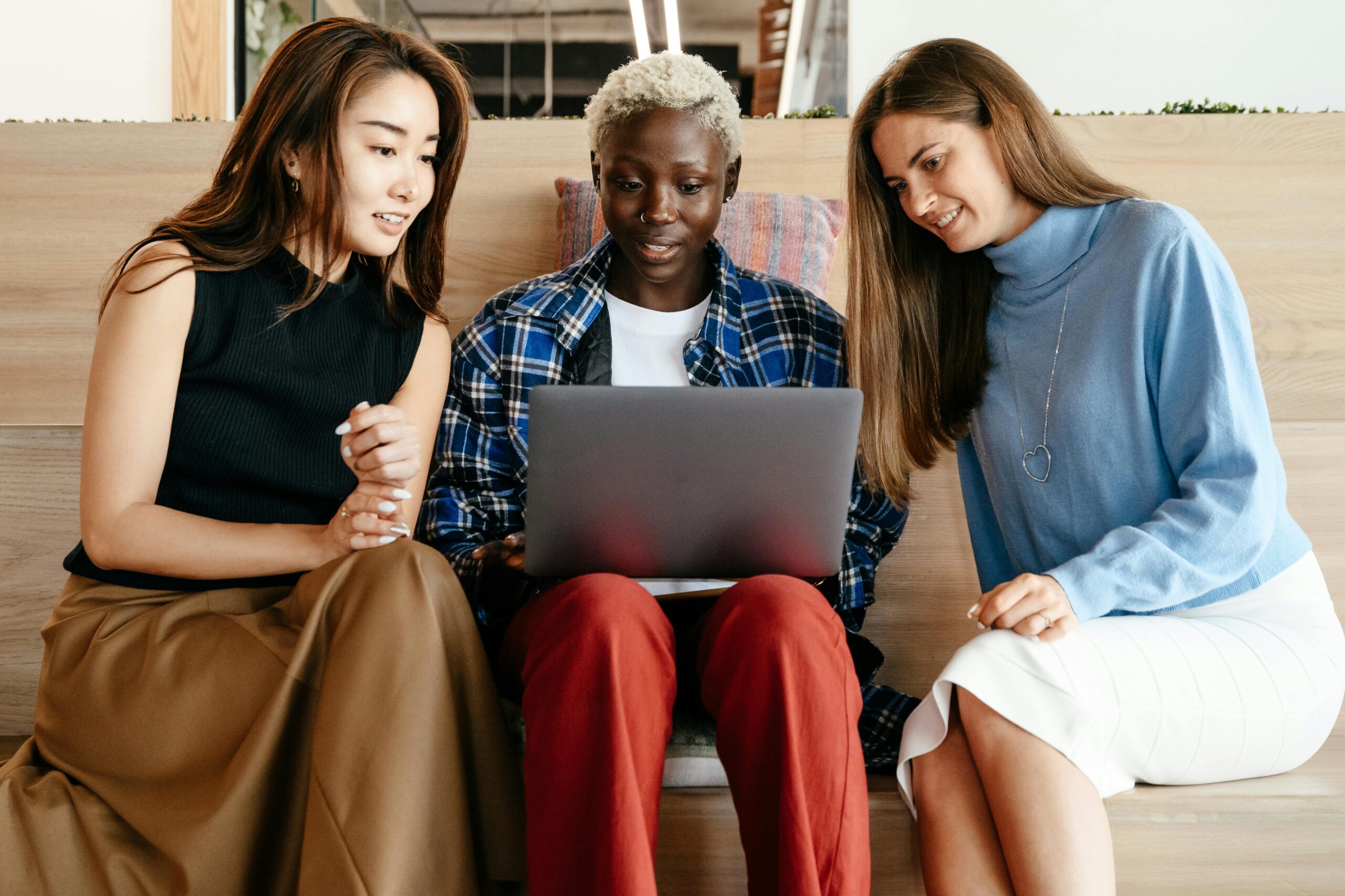 Three diverse young women collaborating on a laptop, indoors, in a modern workspace.