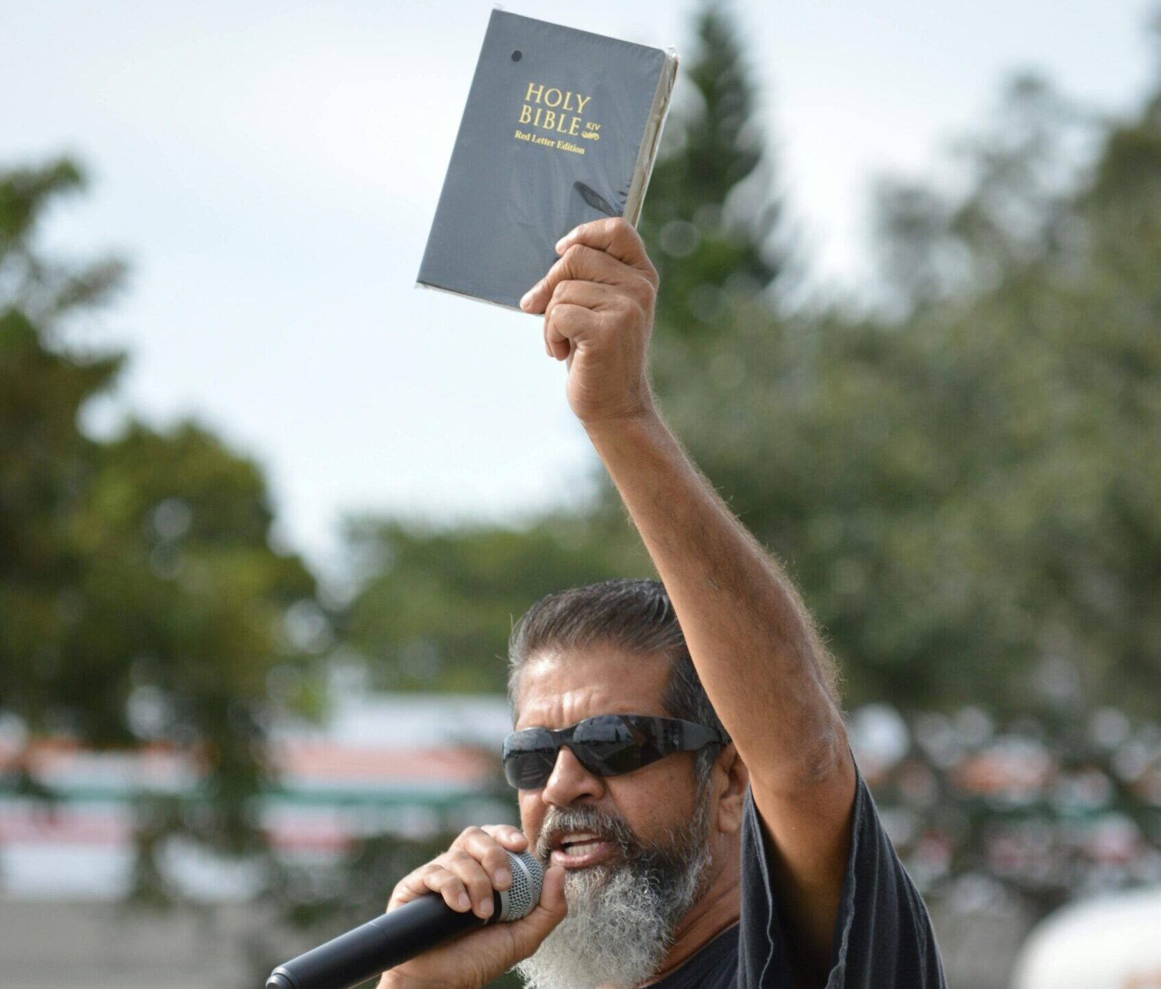 Man with microphone and Bible speaking passionately outdoors.